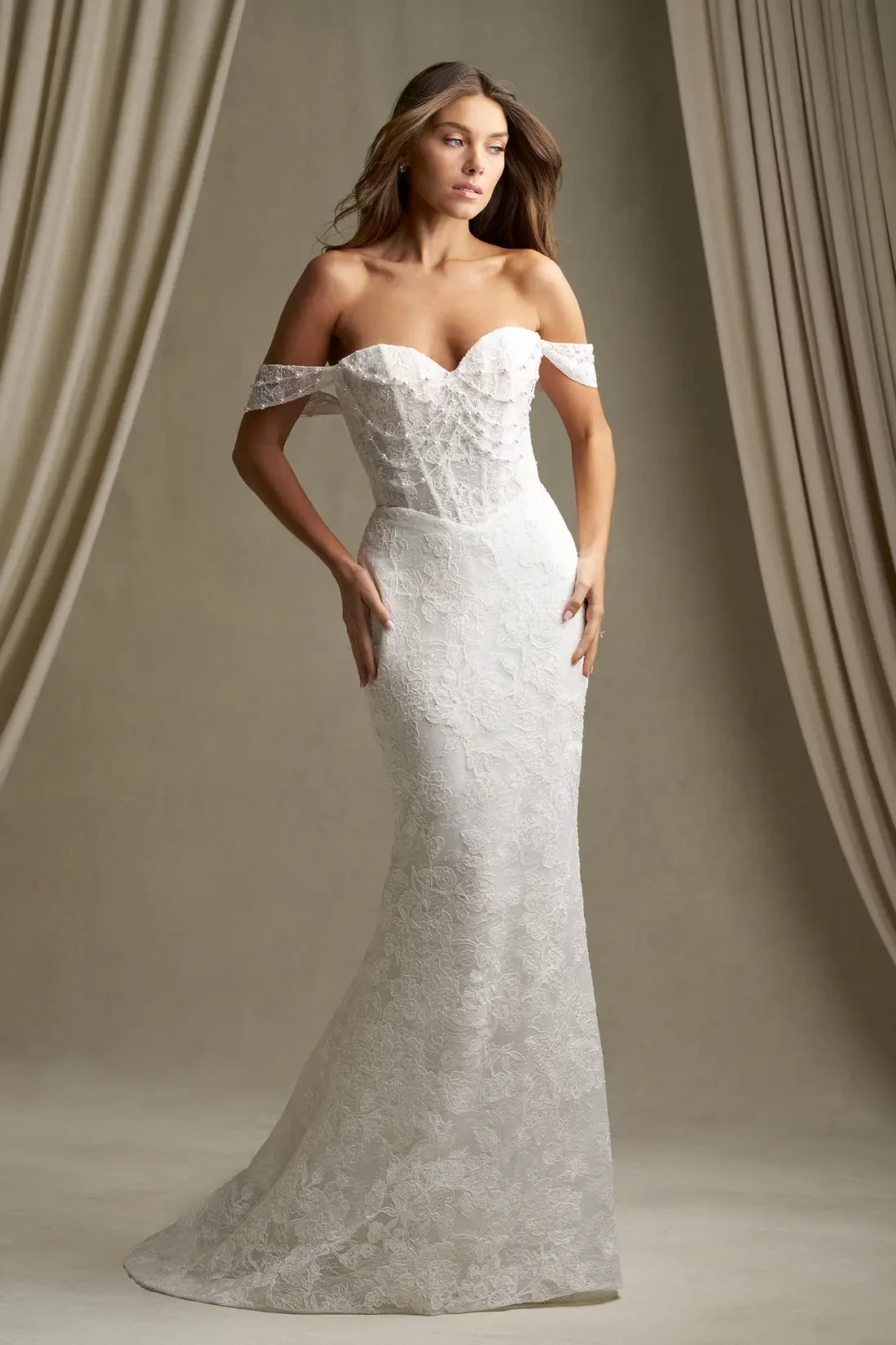 A model is wearing an elegant white off-shoulder wedding gown with lace details, posing in front of soft draped fabric.
