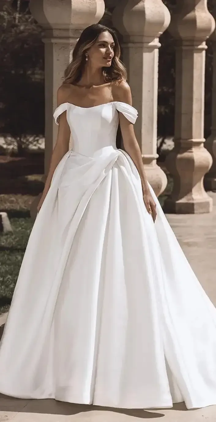 A woman in a white off-shoulder wedding gown with a large bow at the waist, standing outdoors near pillars.