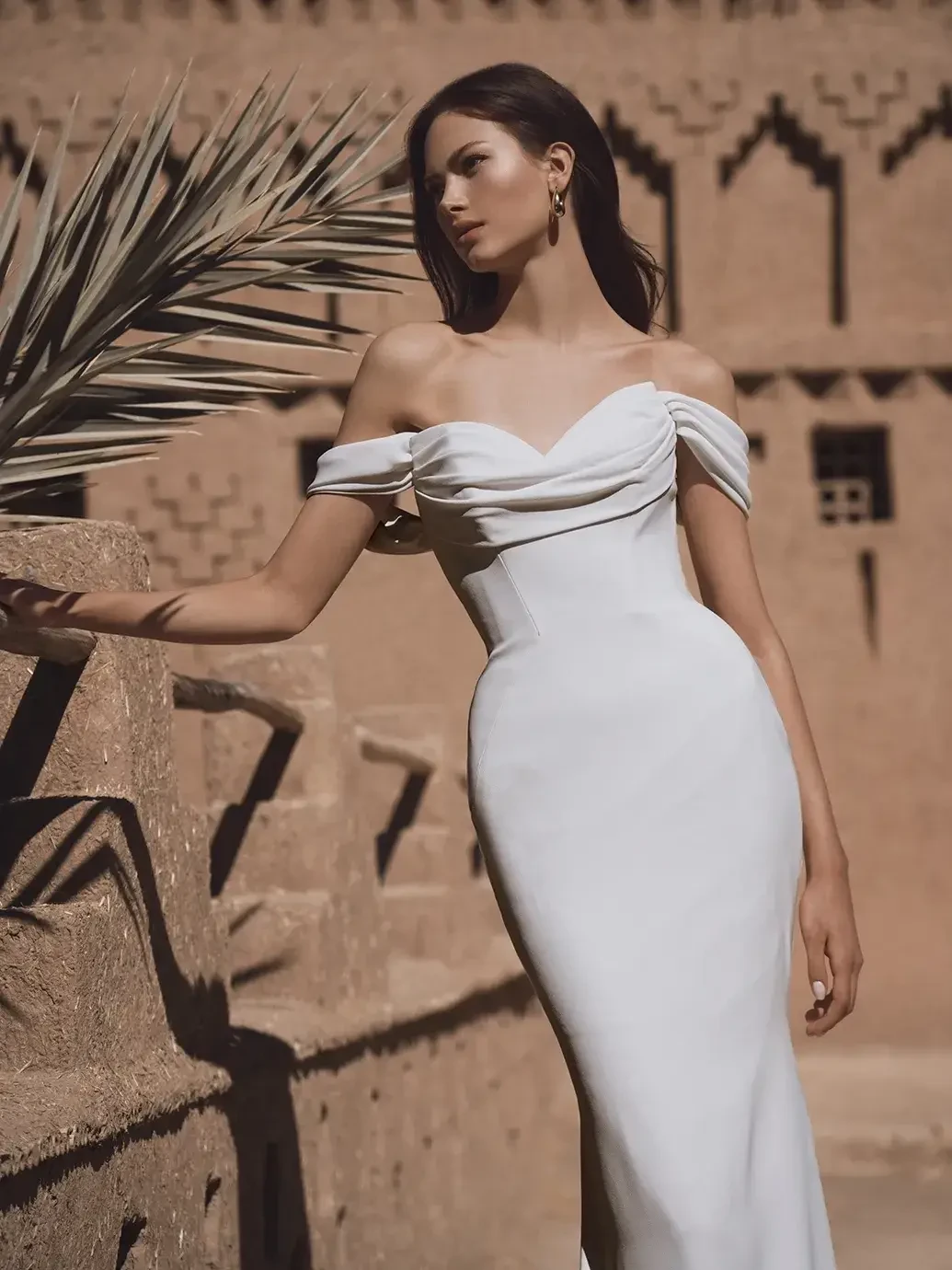 A woman wearing a fitted white dress with off-the-shoulder sleeves stands against a textured background, featuring an architectural design.